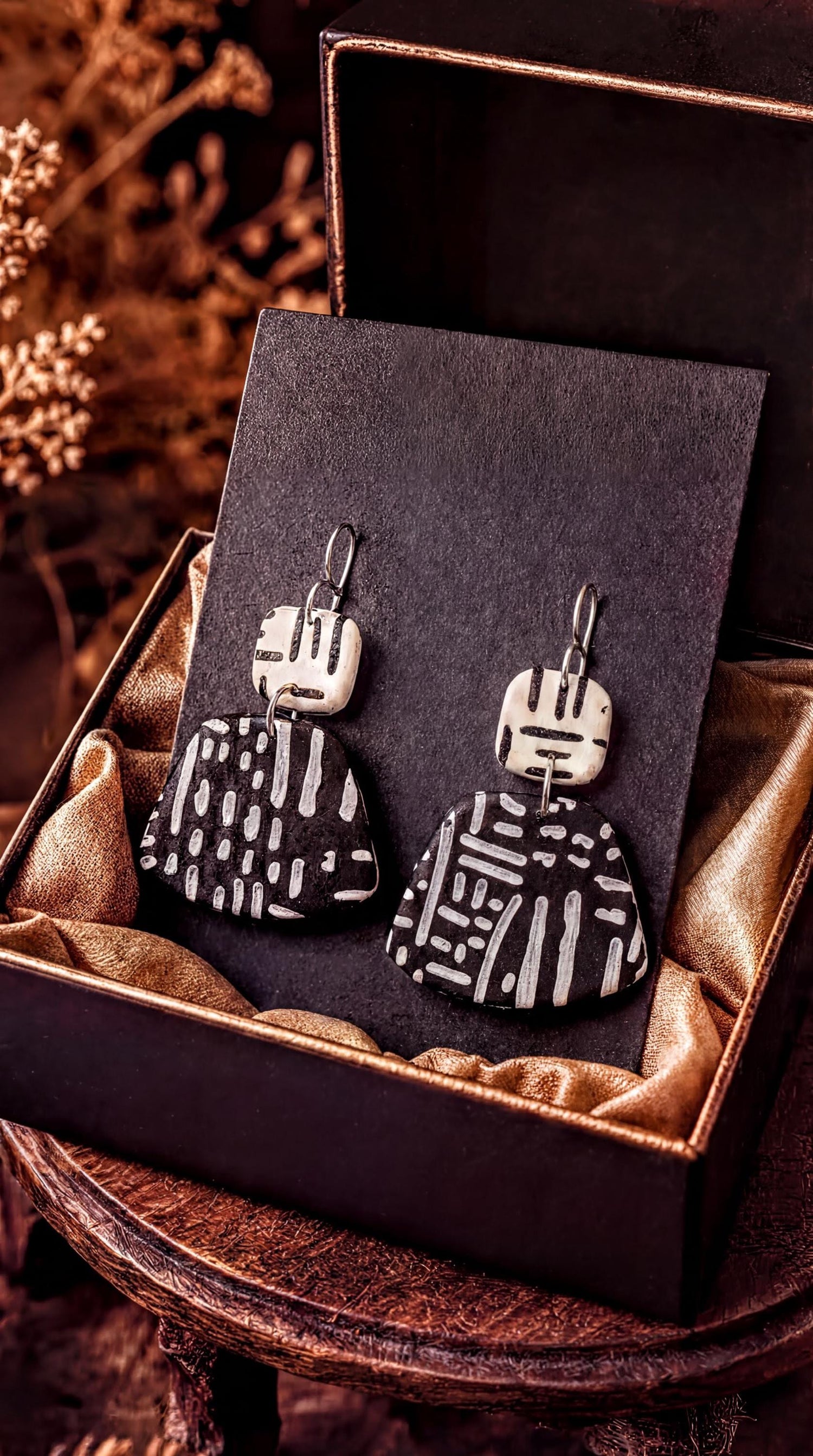 A pair of black and white patterned earrings are displayed in a black box with a gold lining, placed on a wooden surface.