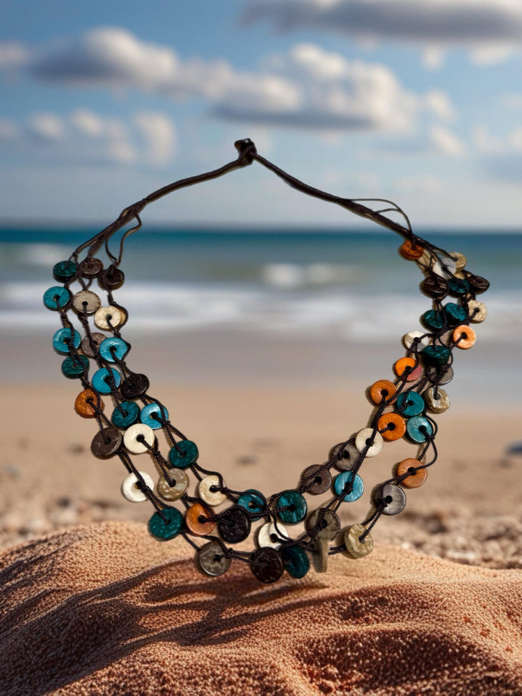 A colorful beaded necklace is displayed on a sandy beach, with the ocean and cloudy sky visible in the background.