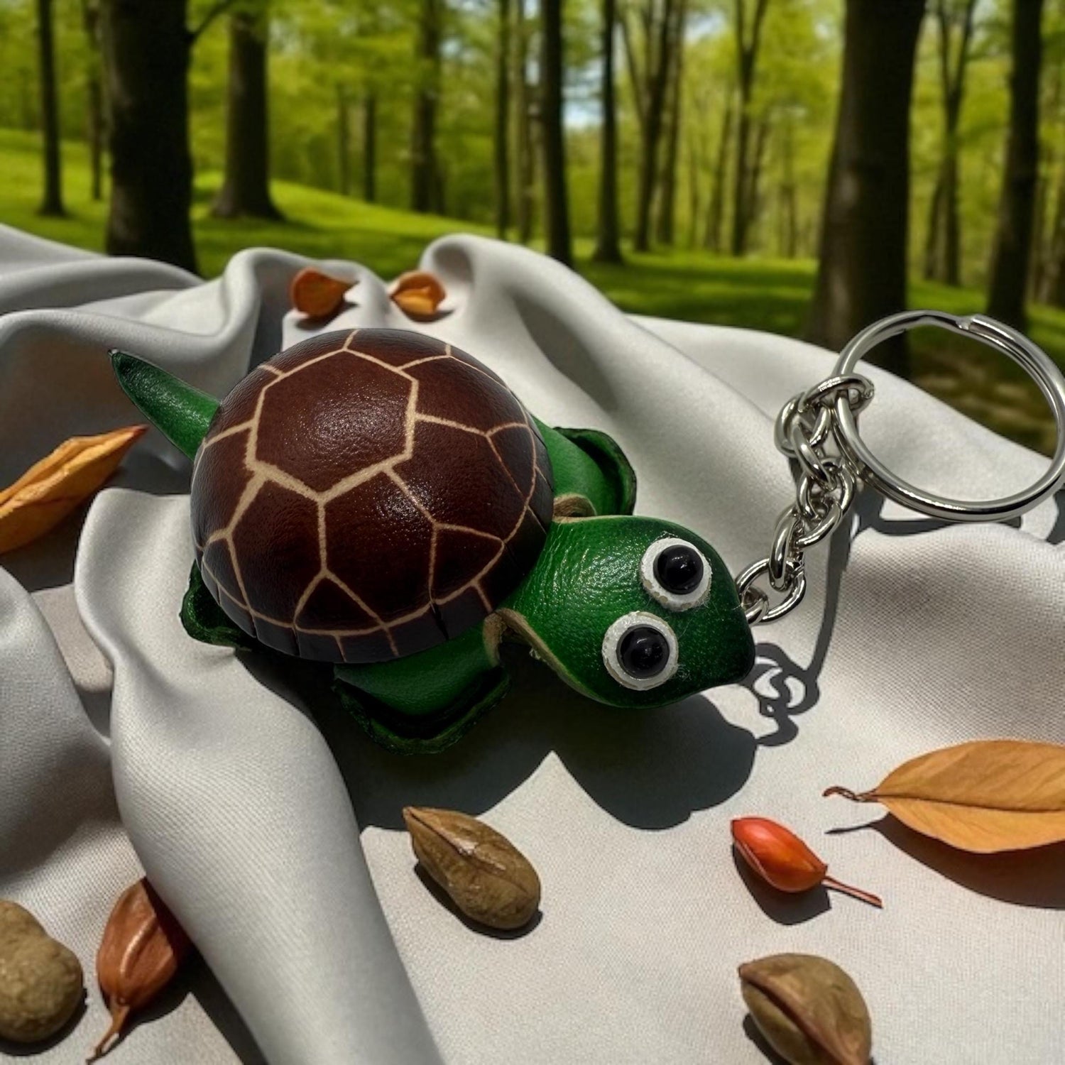 A green turtle keychain with brown spots and two large eyes is placed on a white cloth, surrounded by fallen leaves and small nuts.