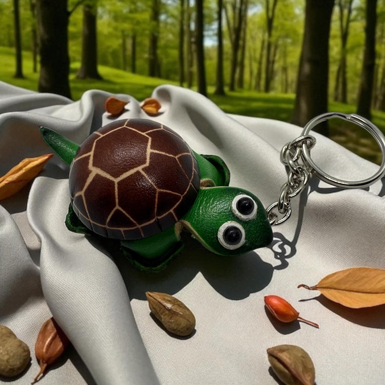 A green turtle keychain with brown spots and two large eyes is placed on a white cloth, surrounded by fallen leaves and small nuts.