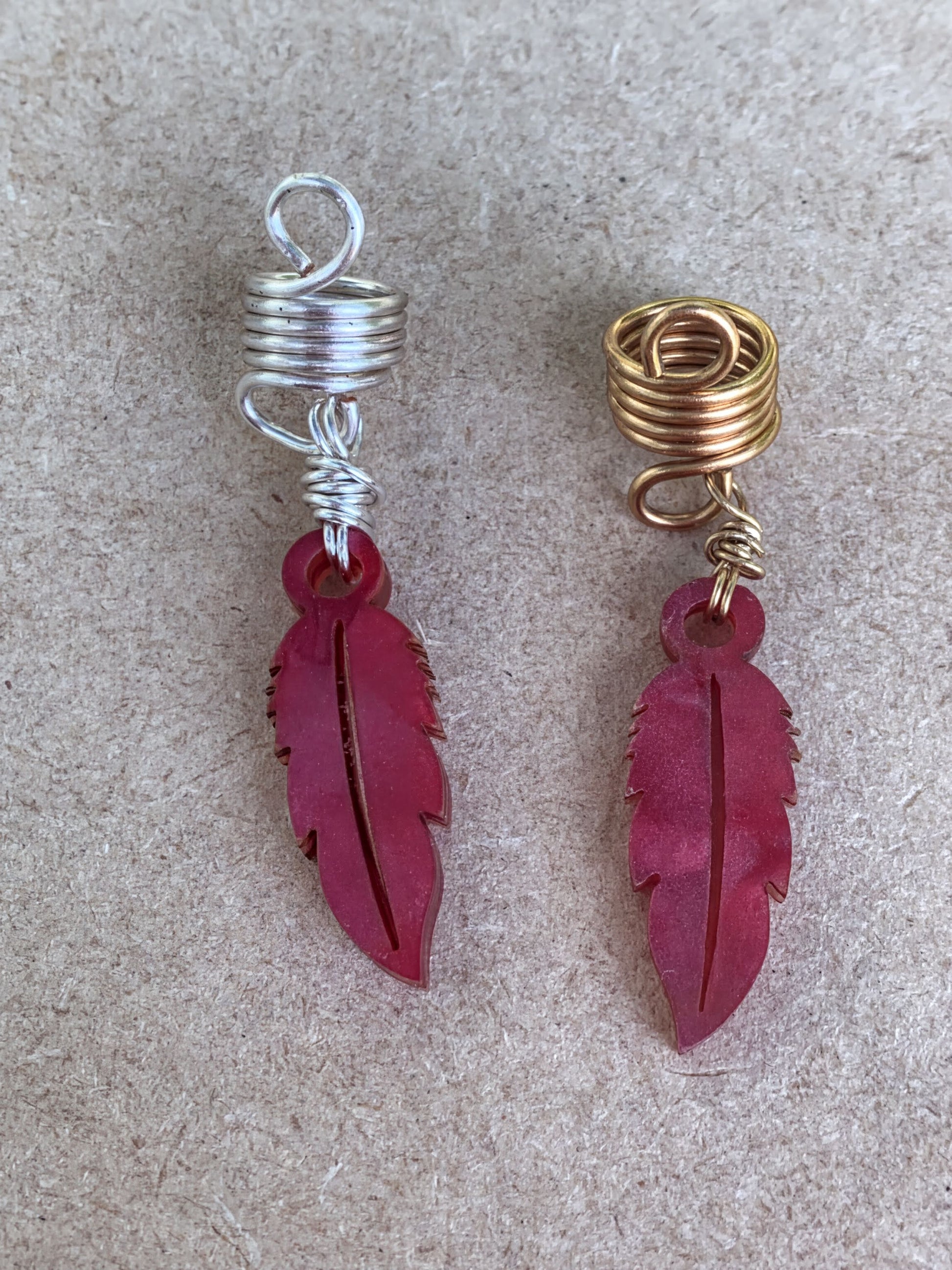 two red feather earrings with spiral designs, one silver and one gold, placed on a textured surface.