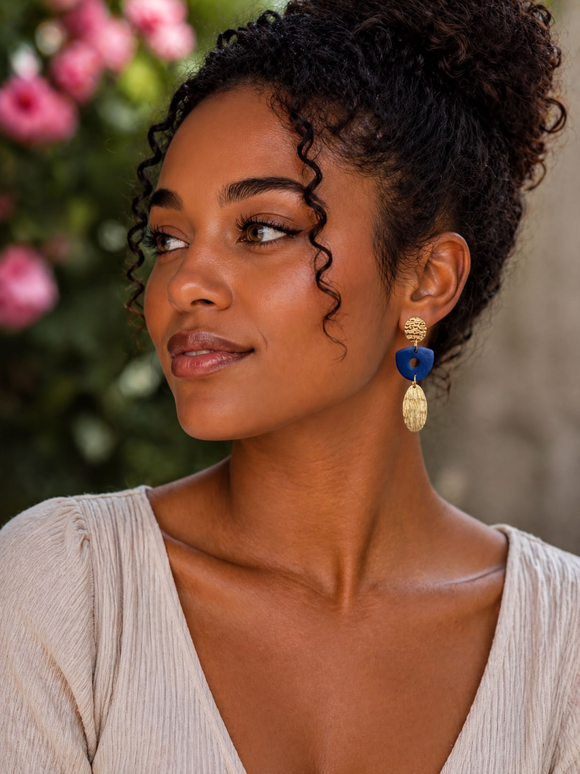 A woman with curly hair wearing a white blouse and gold earrings with blue accents, looking off to the side with a thoughtful expression.