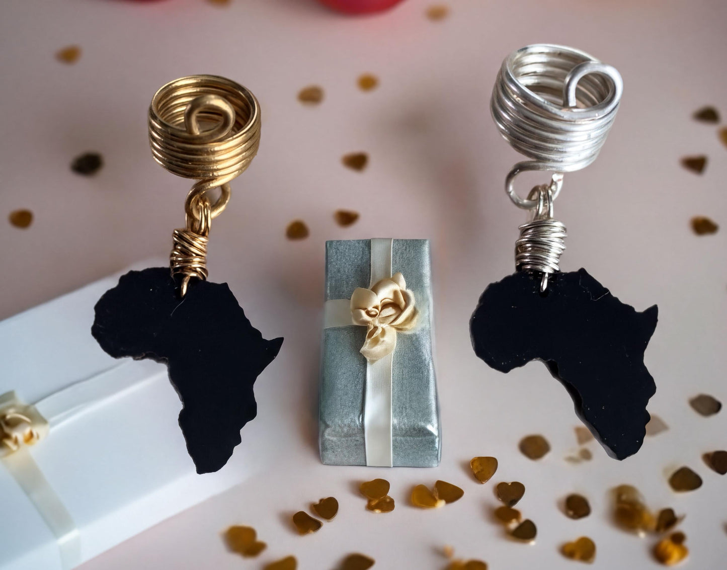 a pair of earrings with African continent designs, a small gift box with a gold ribbon, and scattered gold confetti on a pink background.