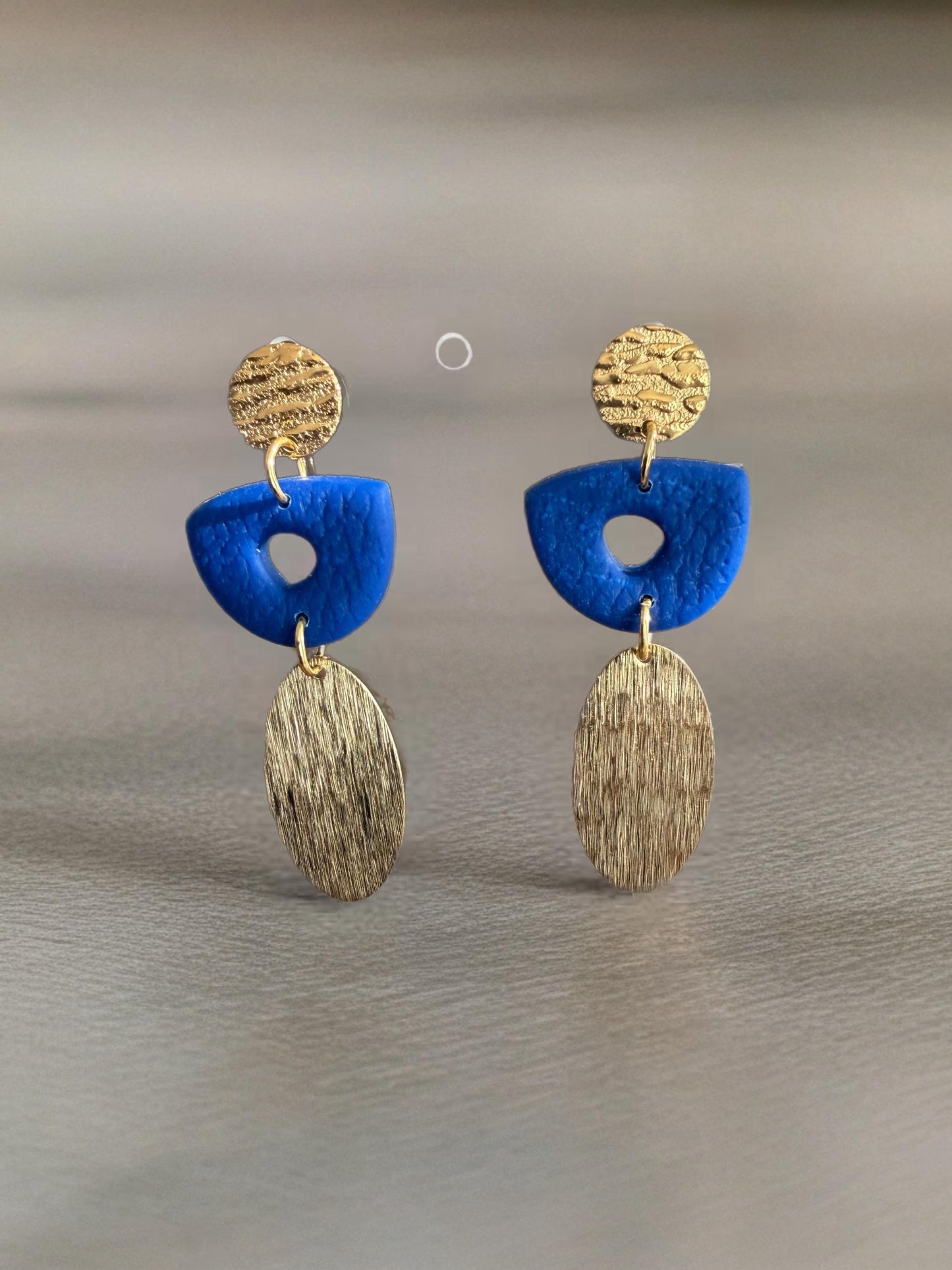 A pair of earrings with blue and gold elements, including a blue oval-shaped piece and a gold circular piece with a textured surface.