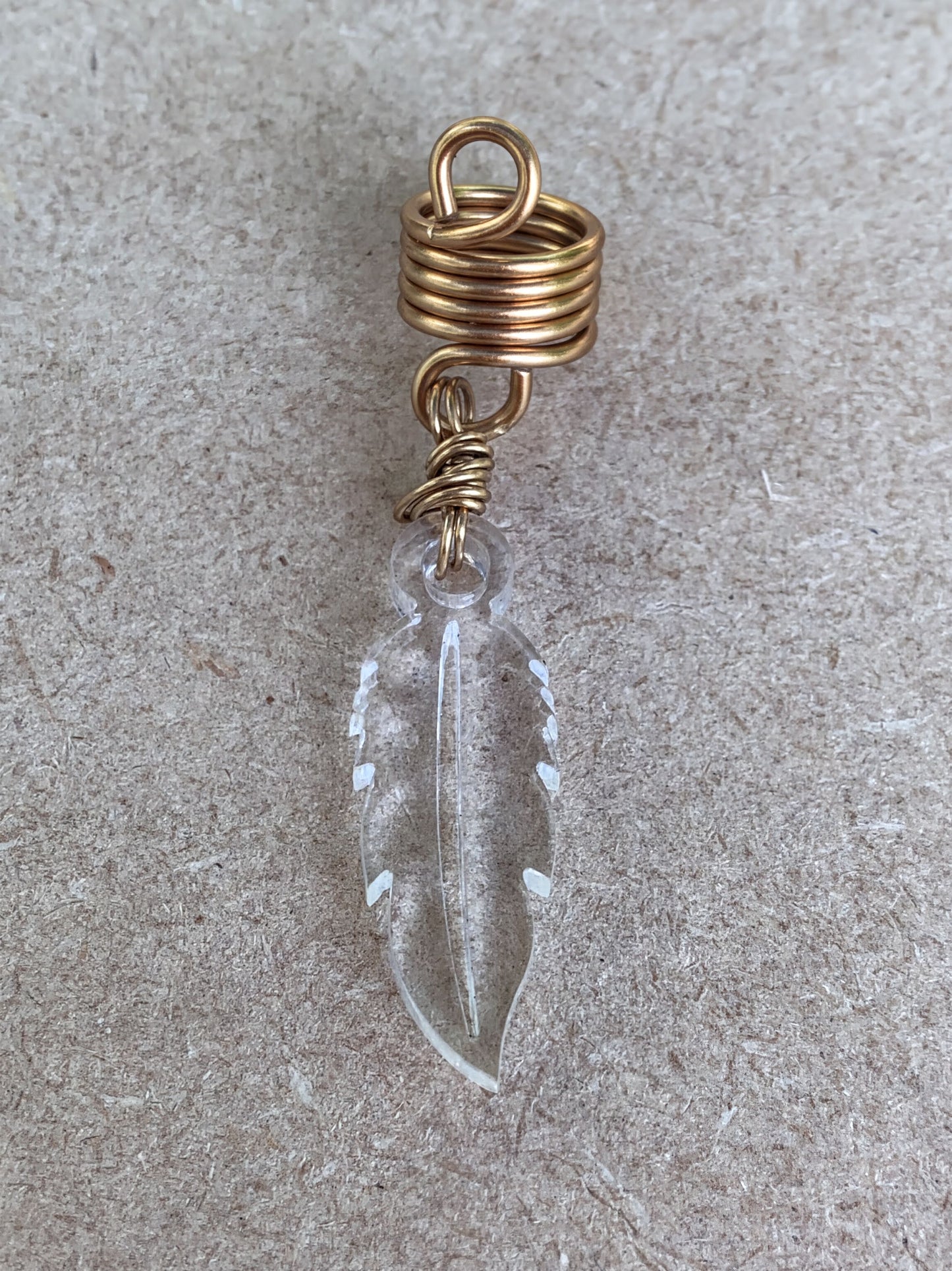 A clear glass feather earring with a gold spiral attached to it, resting on a textured surface.