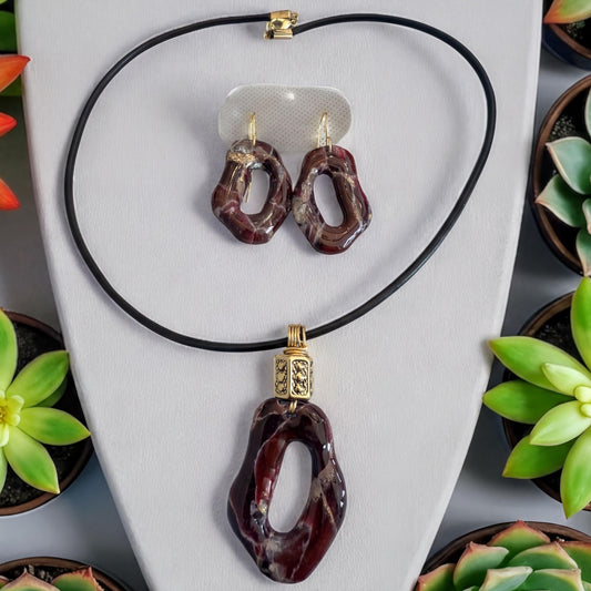 a necklace, earrings, and a pendant set made of a dark-colored stone or gemstone, possibly a type of marble or jade. The necklace and earrings are hanging from a black cord, and the pendant is a large, circular piece with a unique design. The background features several potted plants, creating a natural and decorative setting for the jewelry display.