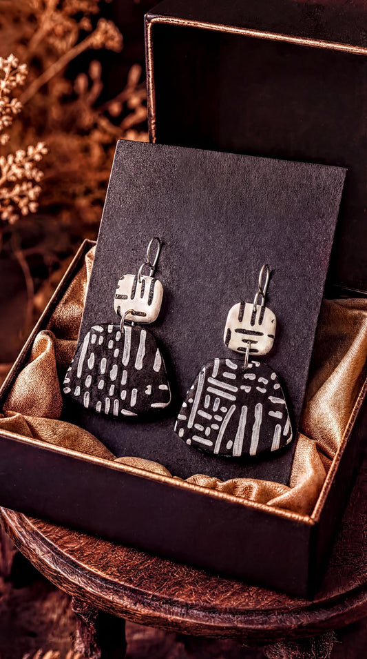 A pair of black and white patterned earrings are displayed in a black box with a gold lining, placed on a wooden surface.