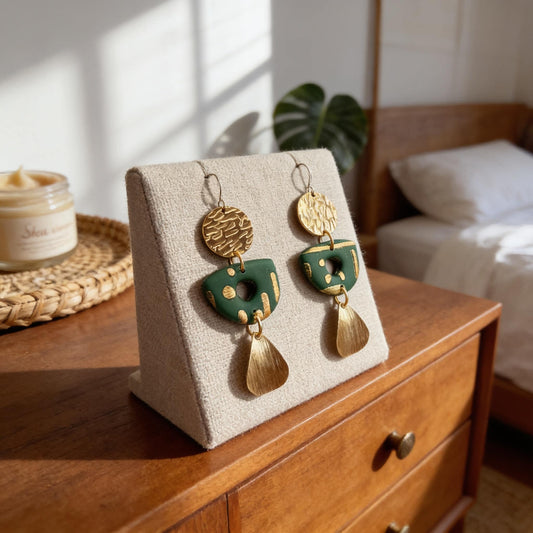 A pair of green and gold earrings displayed on a wooden stand, with a candle and a plant in the background.