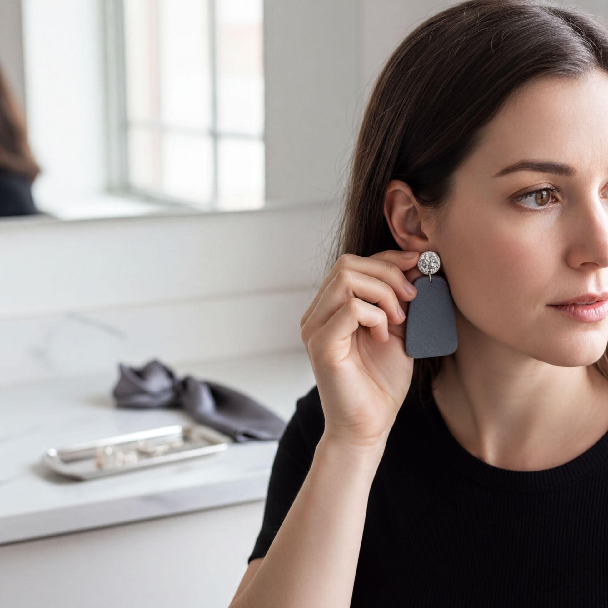 A woman is adjusting a pair of gray earrings in a bathroom setting, with a mirror and a silver tray visible in the background.