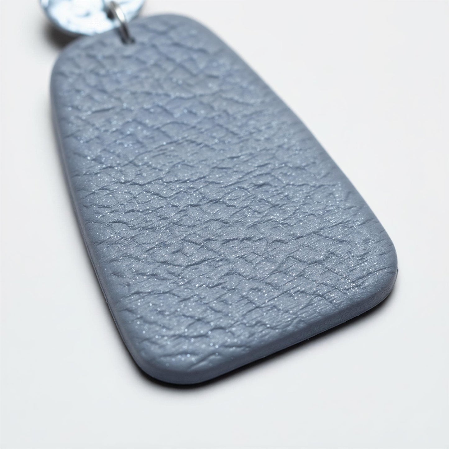 A close-up image of a grey, textured pendant or earring with a metallic or silver-colored metal element attached to it.
