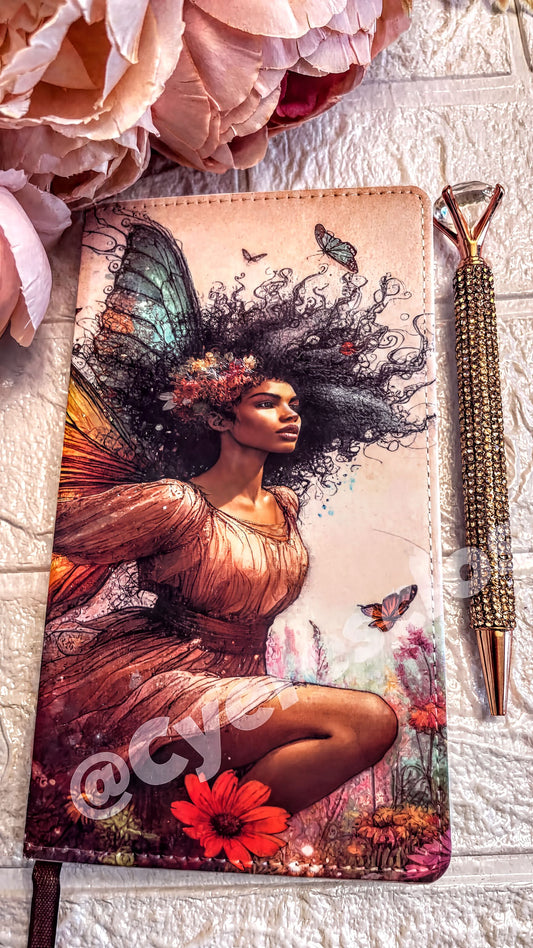 a notebook cover with a woman's face and wings depicted on it, along with flowers and butterflies. The notebook cover is placed on a white surface, and there is a pen next to it.