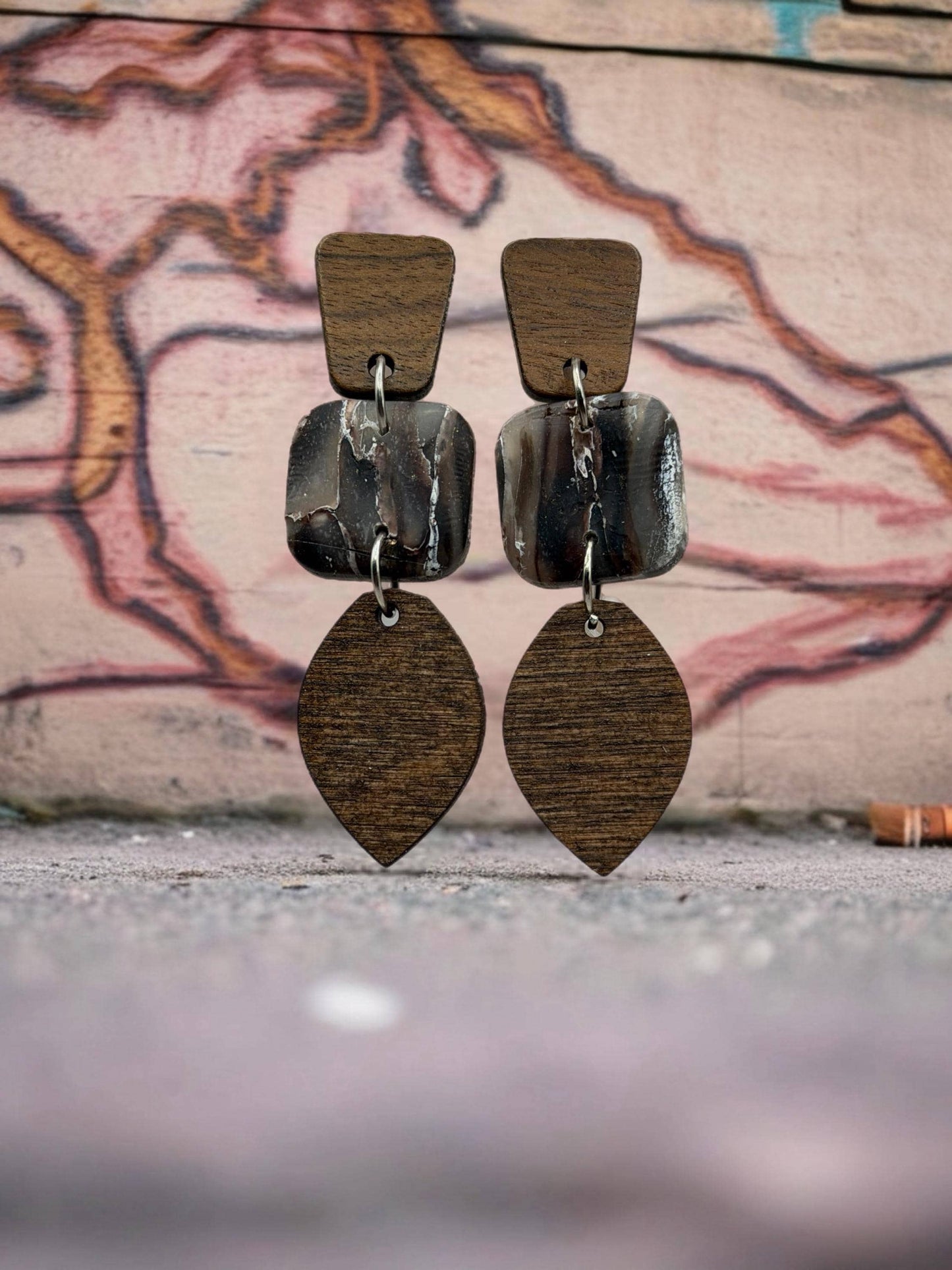 A pair of wooden earrings with a leaf-shaped pendant and a square-shaped pendant, displayed against a graffiti-covered wall.