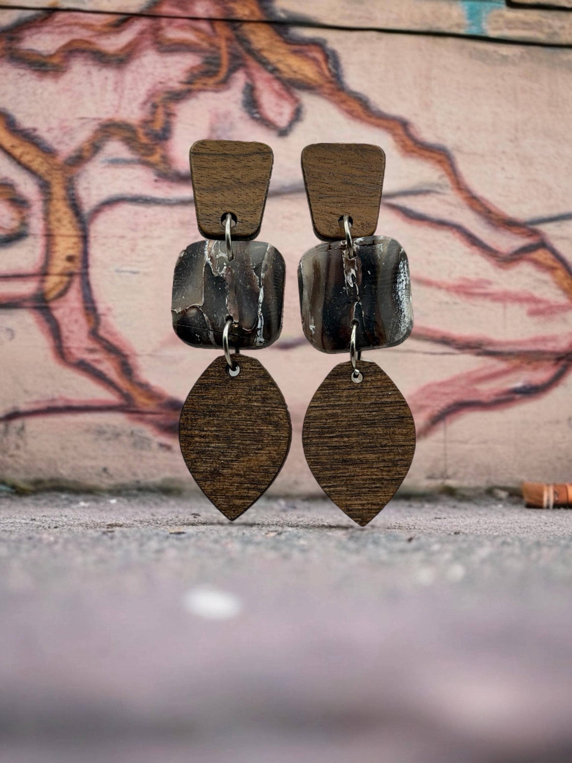 A pair of wooden earrings with a leaf-shaped pendant and a square-shaped pendant, displayed against a graffiti-covered wall.