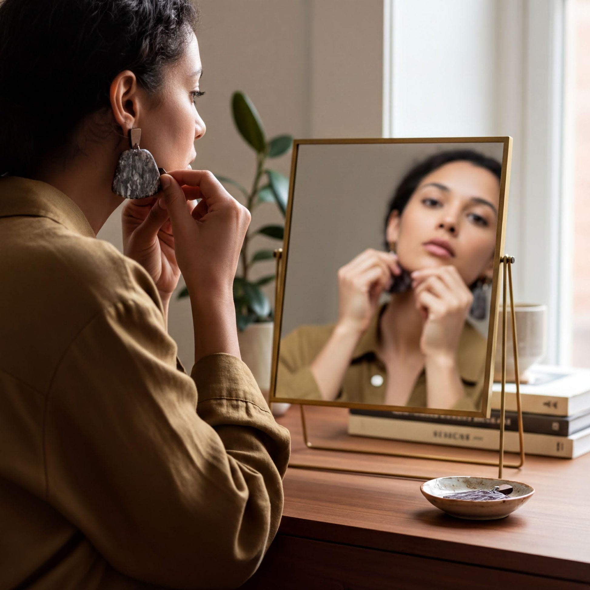 A woman is sitting at a desk, putting on earrings in front of a mirror.