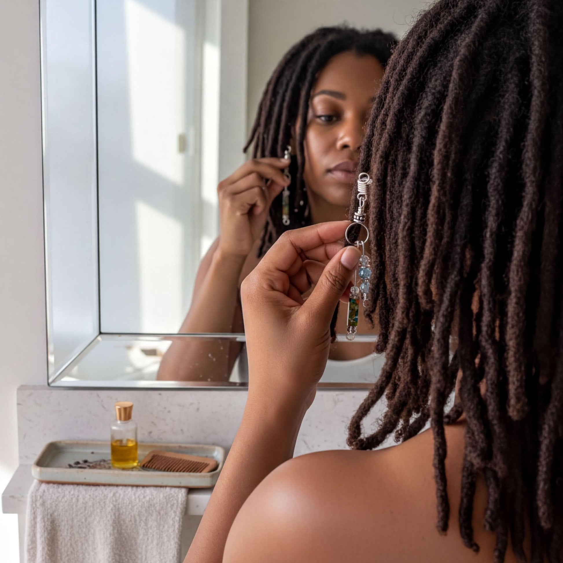 A woman with dreadlocks is standing in front of a mirror, holding a pair of earrings and looking at her reflection.