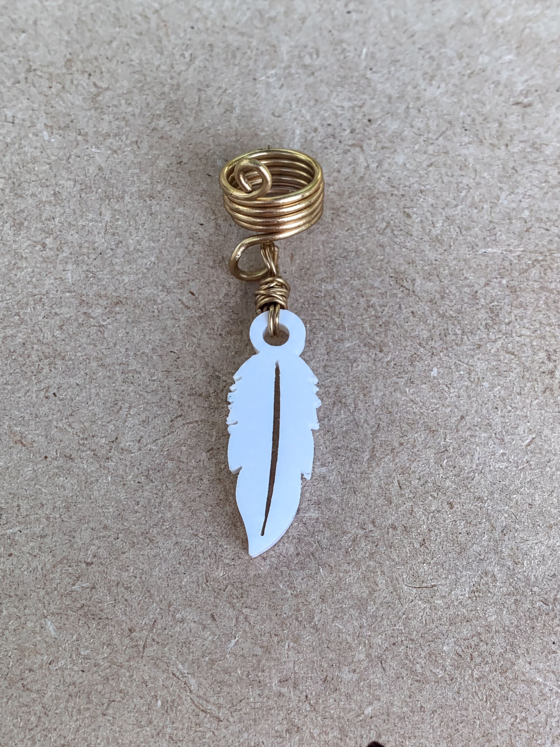 A white feather-shaped earring with a gold spiral attached to it, resting on a textured surface.
