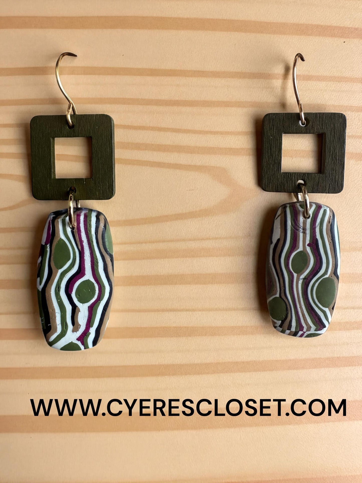 a pair of earrings with a unique, abstract design featuring green and purple swirls and dots. The earrings are hanging from hooks on a wooden surface.