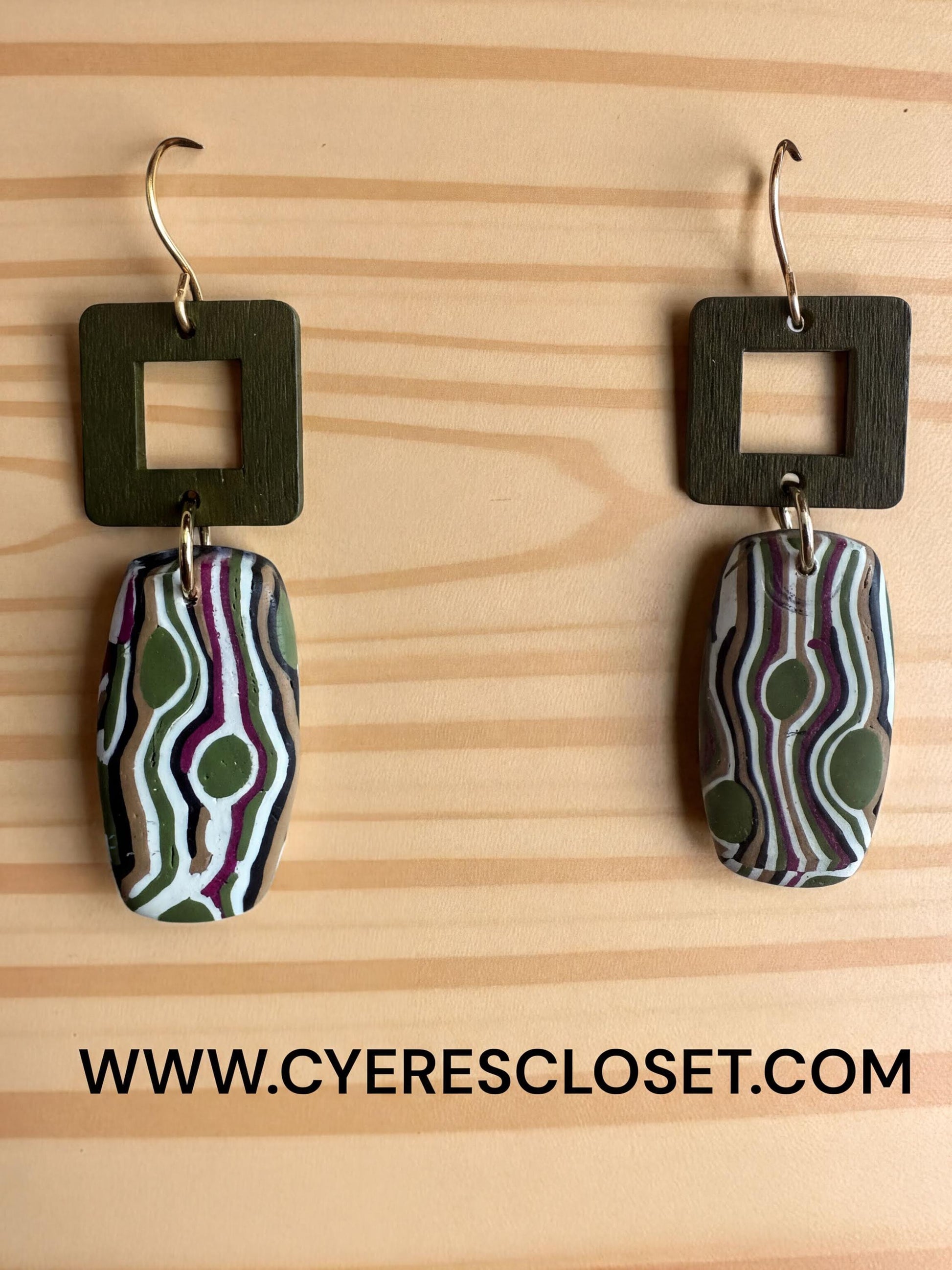 a pair of earrings with a unique, abstract design featuring green and purple swirls and dots. The earrings are hanging from hooks on a wooden surface.