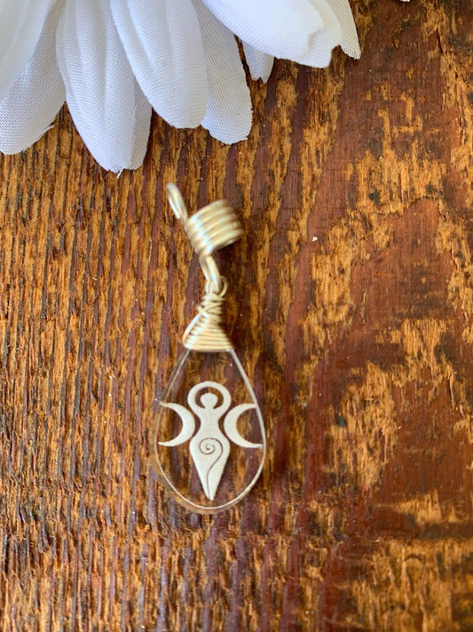 A pendant with a design of a woman with three tails is displayed on a wooden surface, accompanied by a white flower.