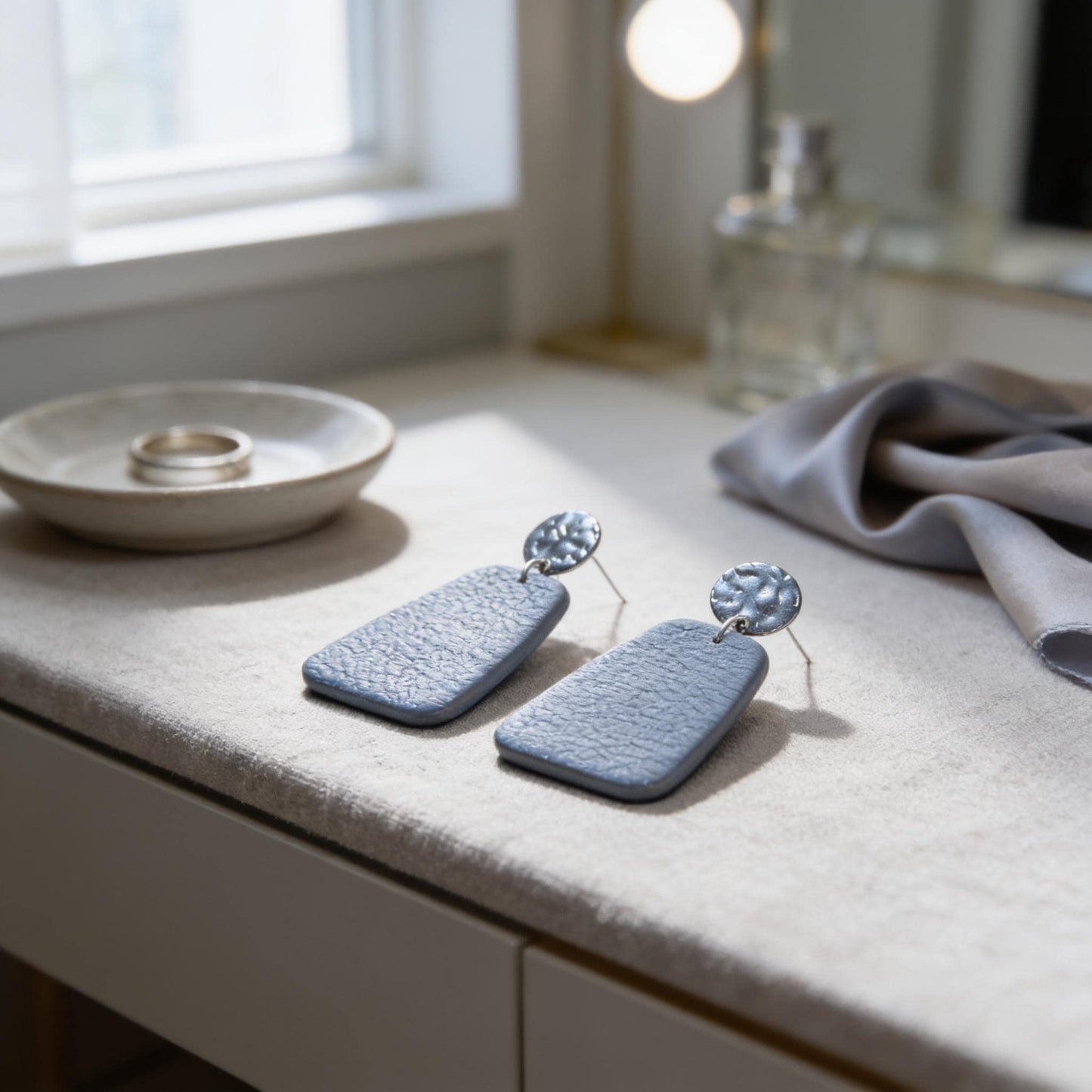 a pair of blue earrings with a silver circular design on the back, placed on a white surface next to a small bowl and a bottle.