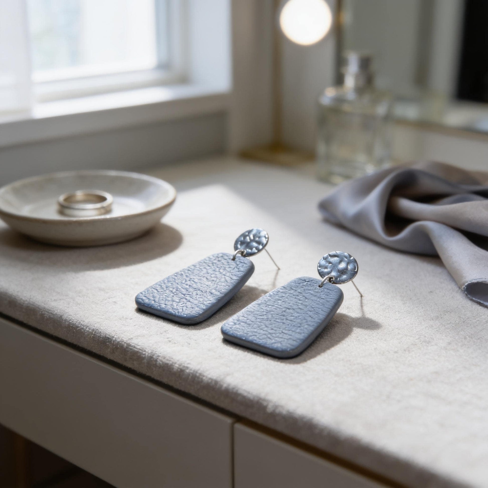 a pair of blue earrings with a silver circular design on the back, placed on a white surface next to a small bowl and a bottle.
