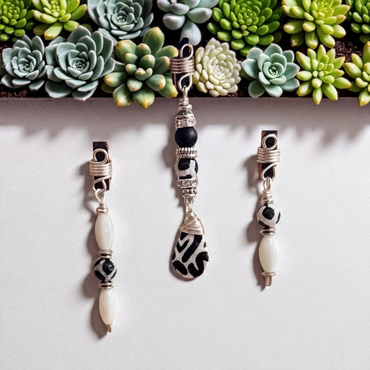 a collection of three unique earrings and pendants, each with distinct designs and materials. The earrings are made of silver wire and have a combination of white and black beads, while the pendants are made of black and white beads. The earrings and pendants are displayed against a white background, with a row of green succulent plants arranged in the top left corner.