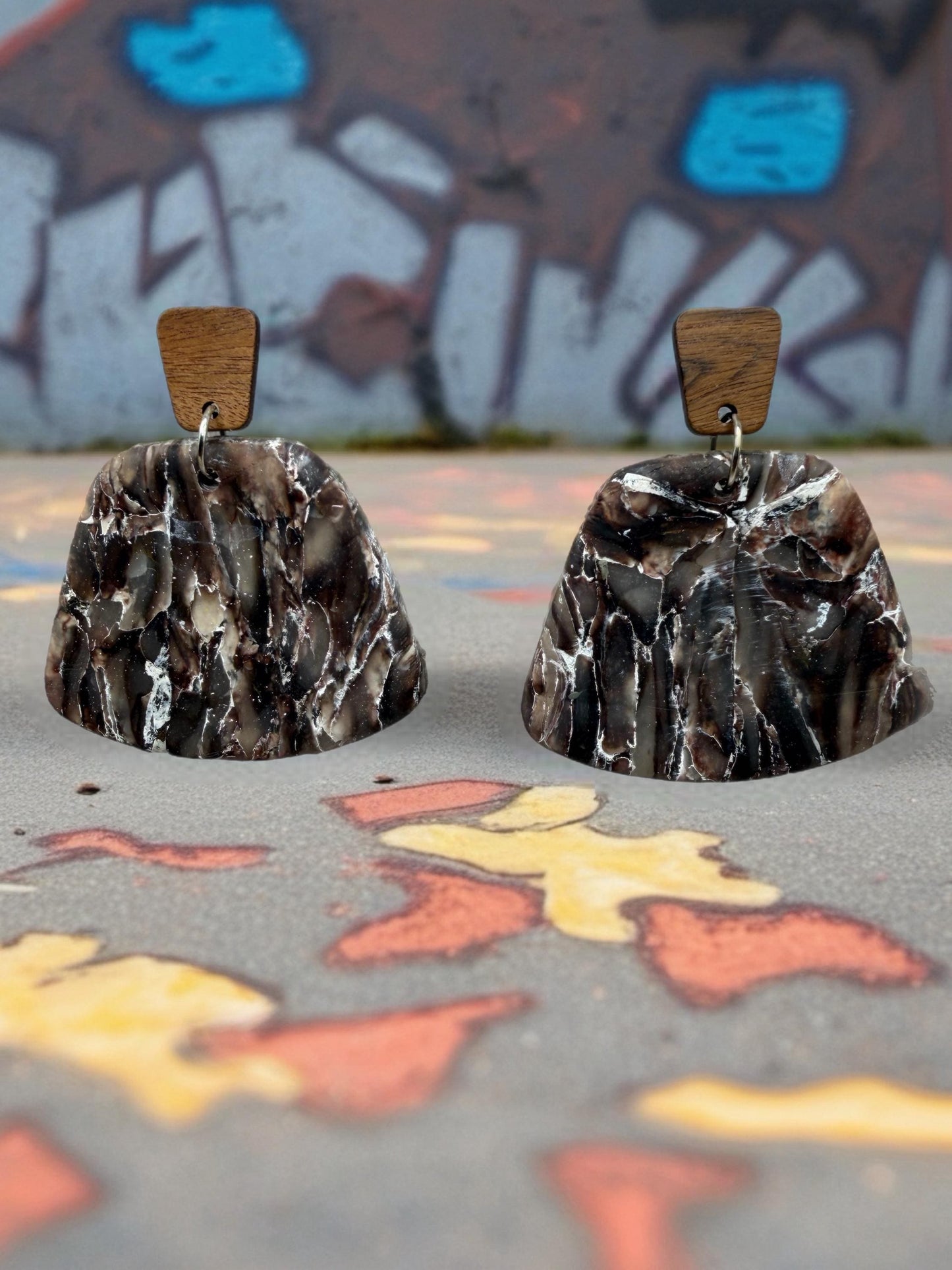 two dark brown, textured earrings with wooden accents, placed on a colorful, graffiti-covered surface.