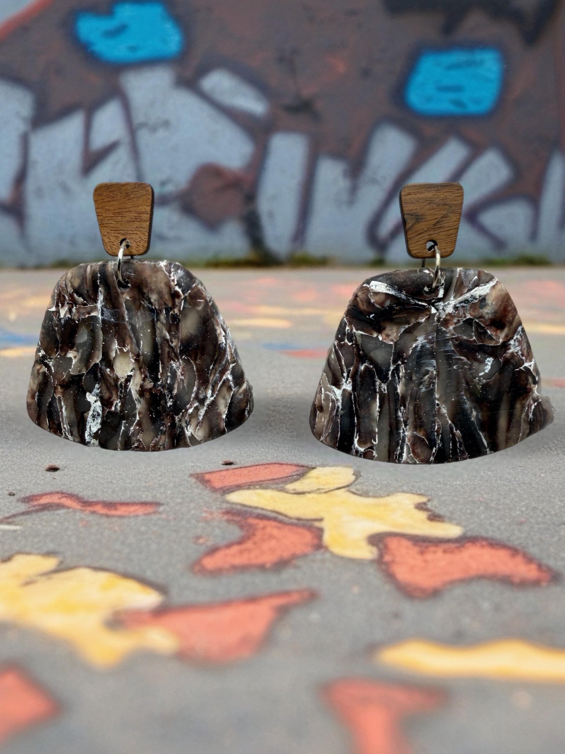 two dark brown, textured earrings with wooden accents, placed on a colorful, graffiti-covered surface.