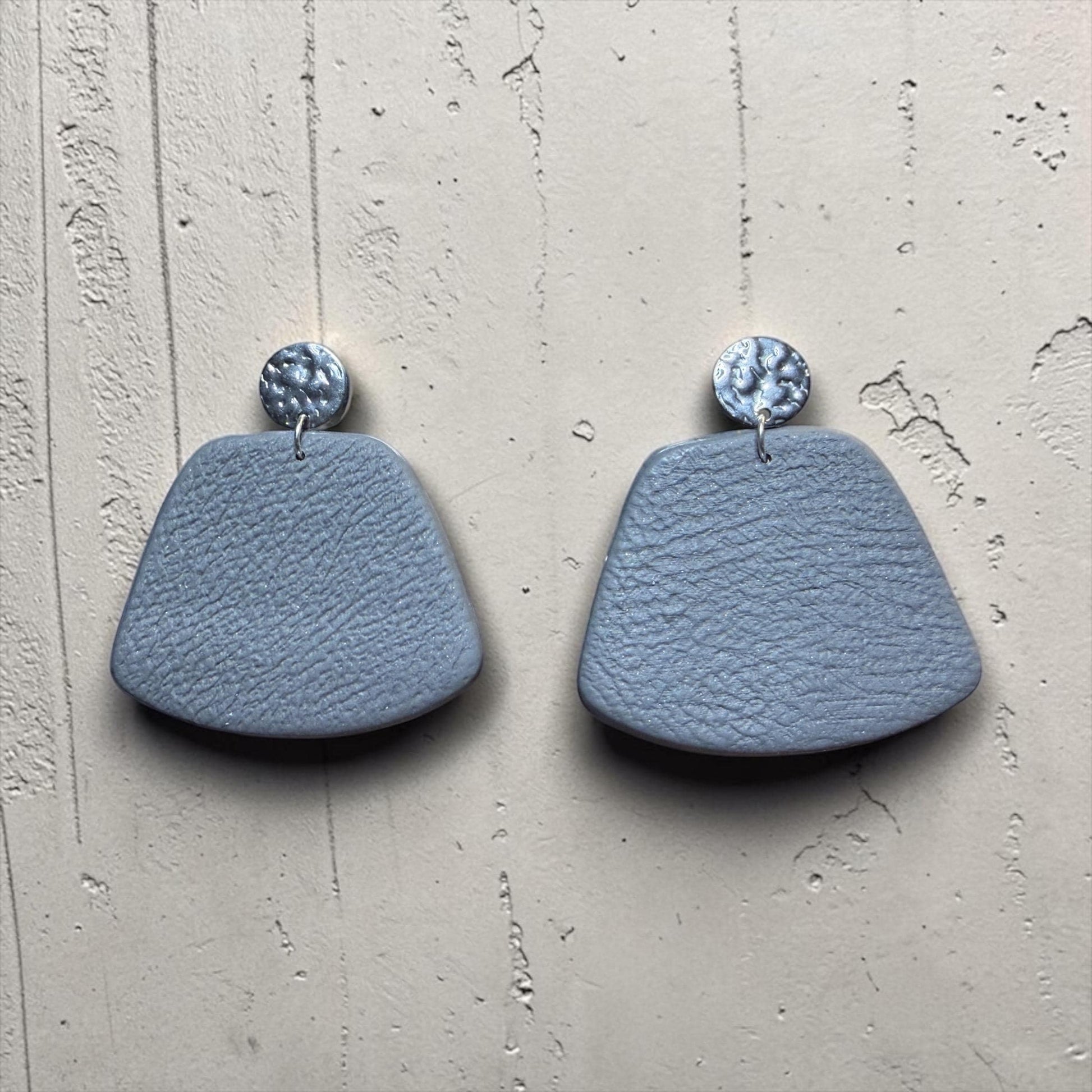 A pair of light blue, textured earrings with small circular charms hanging from them, displayed against a textured white background.