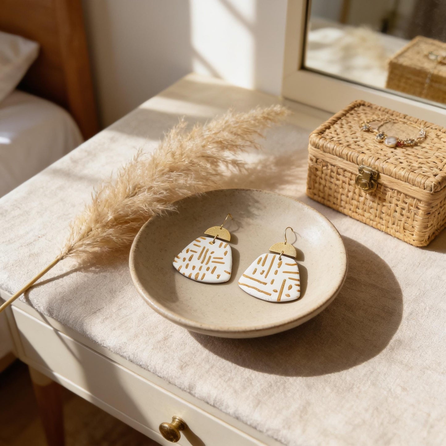 A pair of white earrings with gold accents sits on a white bowl, accompanied by a potted plant and a woven basket on a wooden dresser.