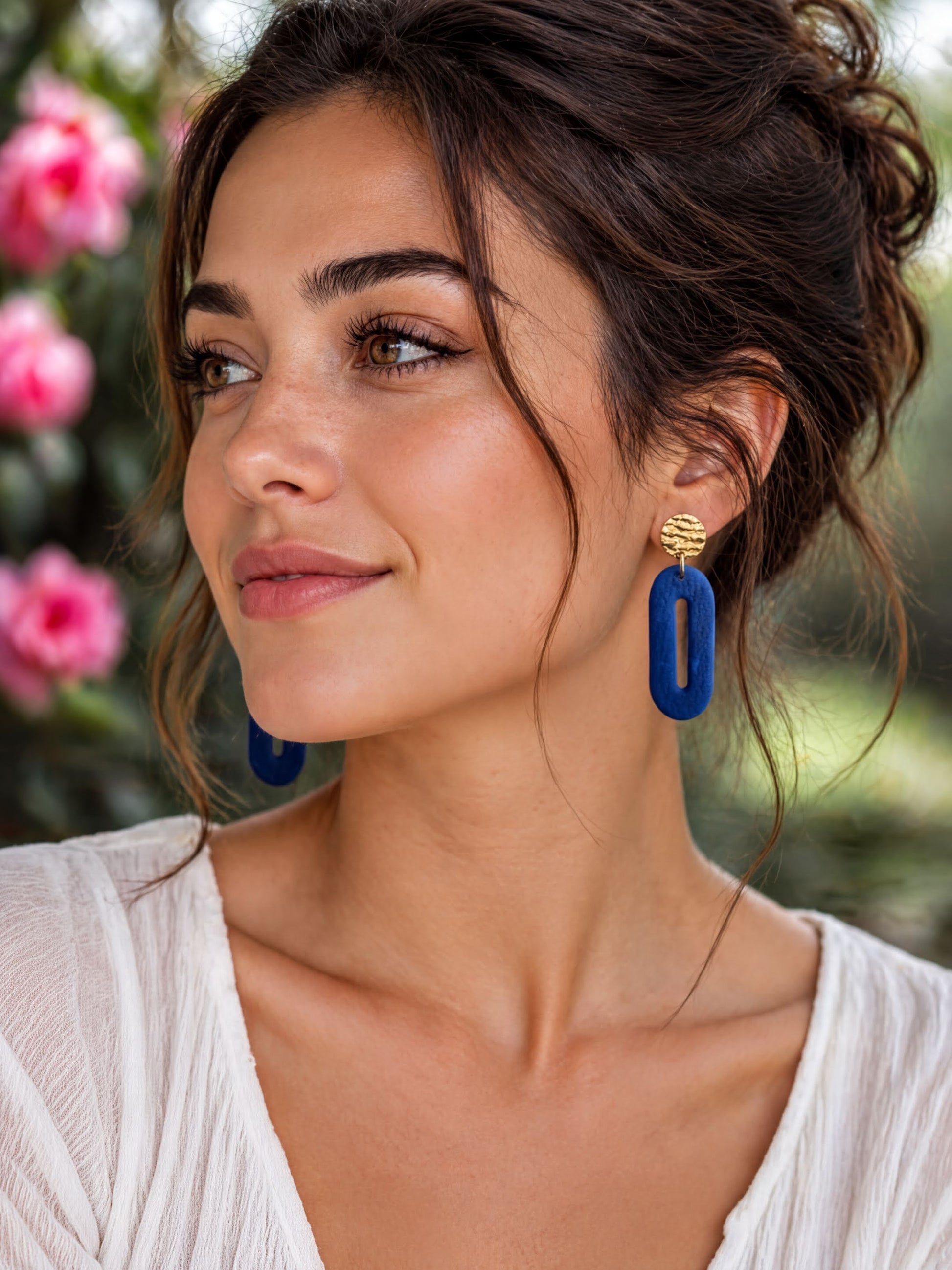 A woman with dark hair and a white blouse is wearing large blue earrings and smiling at the camera.