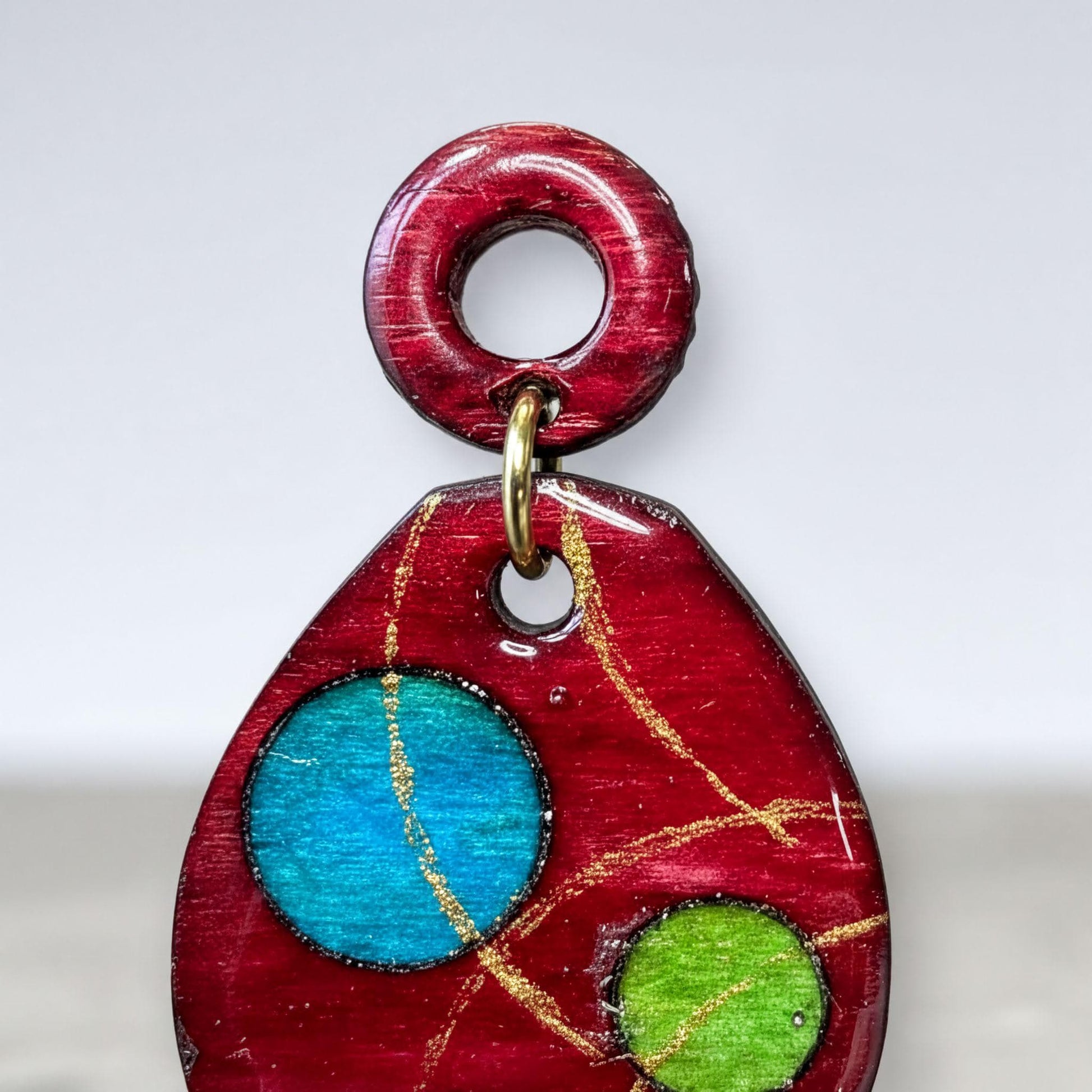 A red pendant with a circular design featuring blue and green elements, and a gold-colored ring hanging from the top.