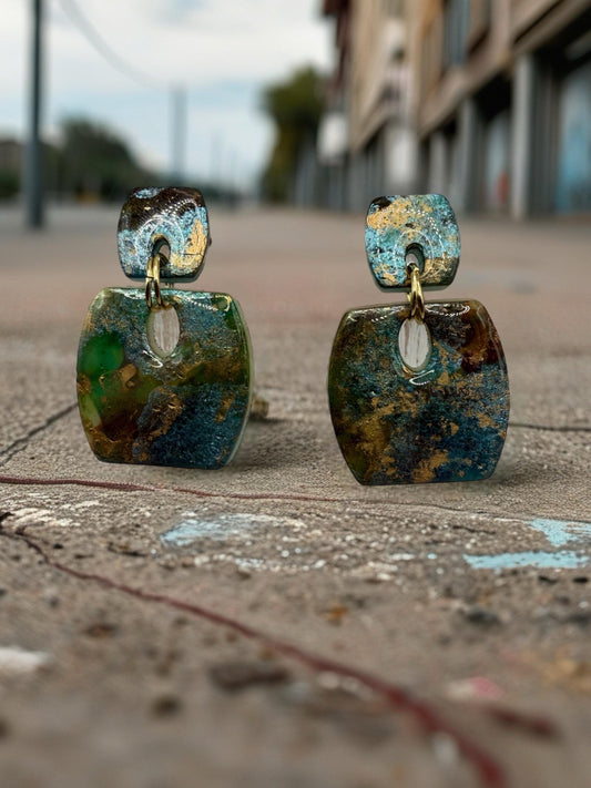 Two square, multicolored earrings with gold-colored accents are displayed on a concrete surface, with a blurred background of buildings and a street.