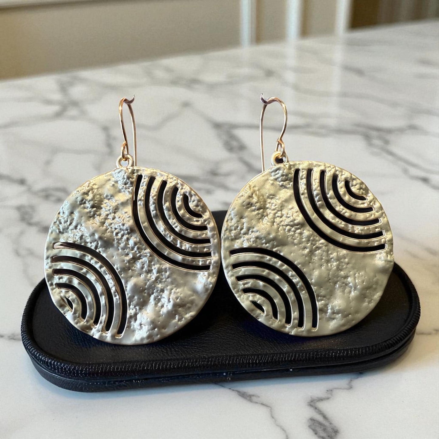 A pair of large, round earrings with a metallic texture and a repeating wave pattern design.
