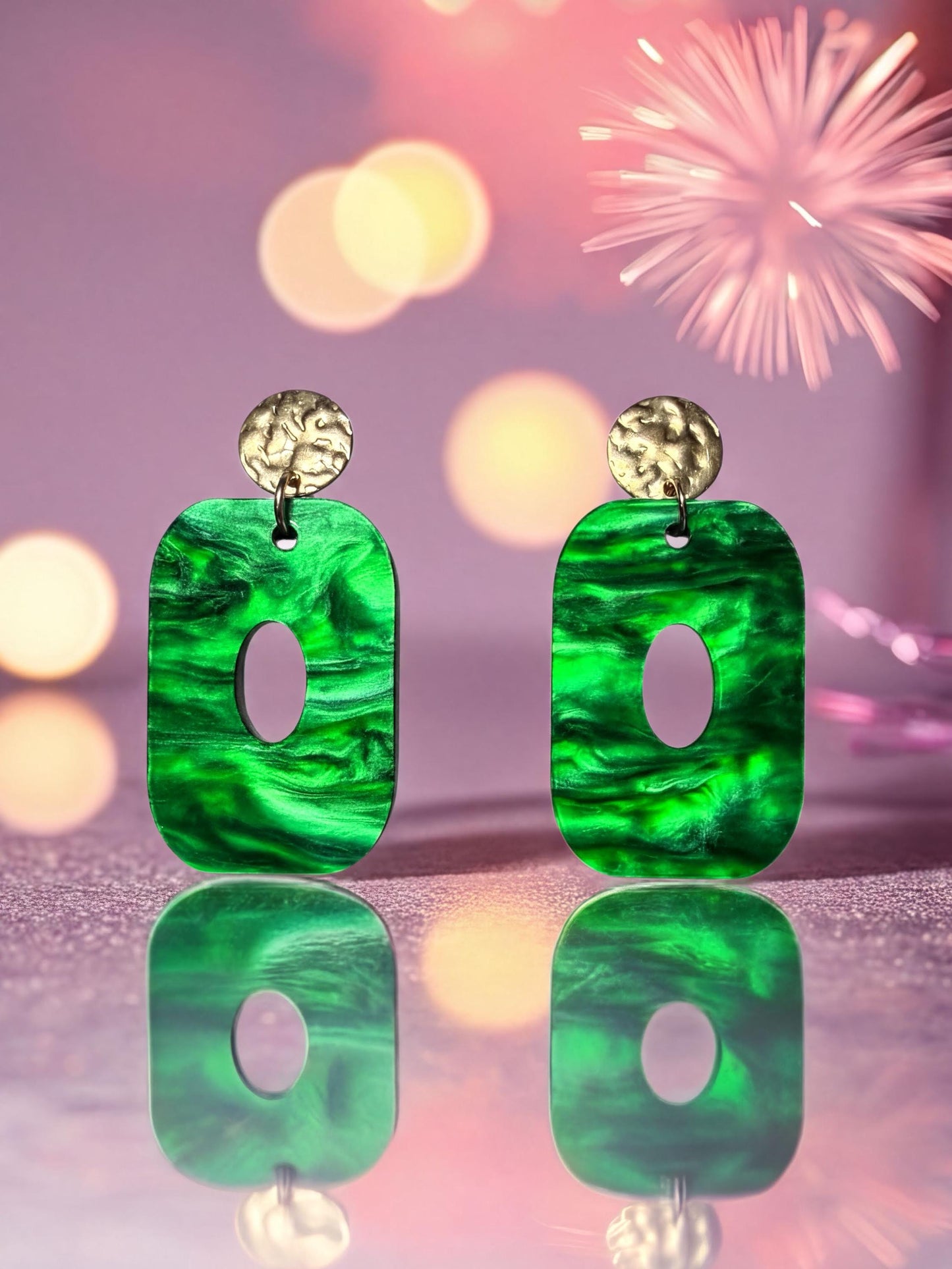 two green earrings with gold accents, set against a background of colorful lights and a sparkly purple surface.