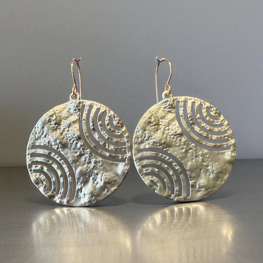 A pair of large, round earrings with a metallic texture and a repeating pattern of waves or circles.
