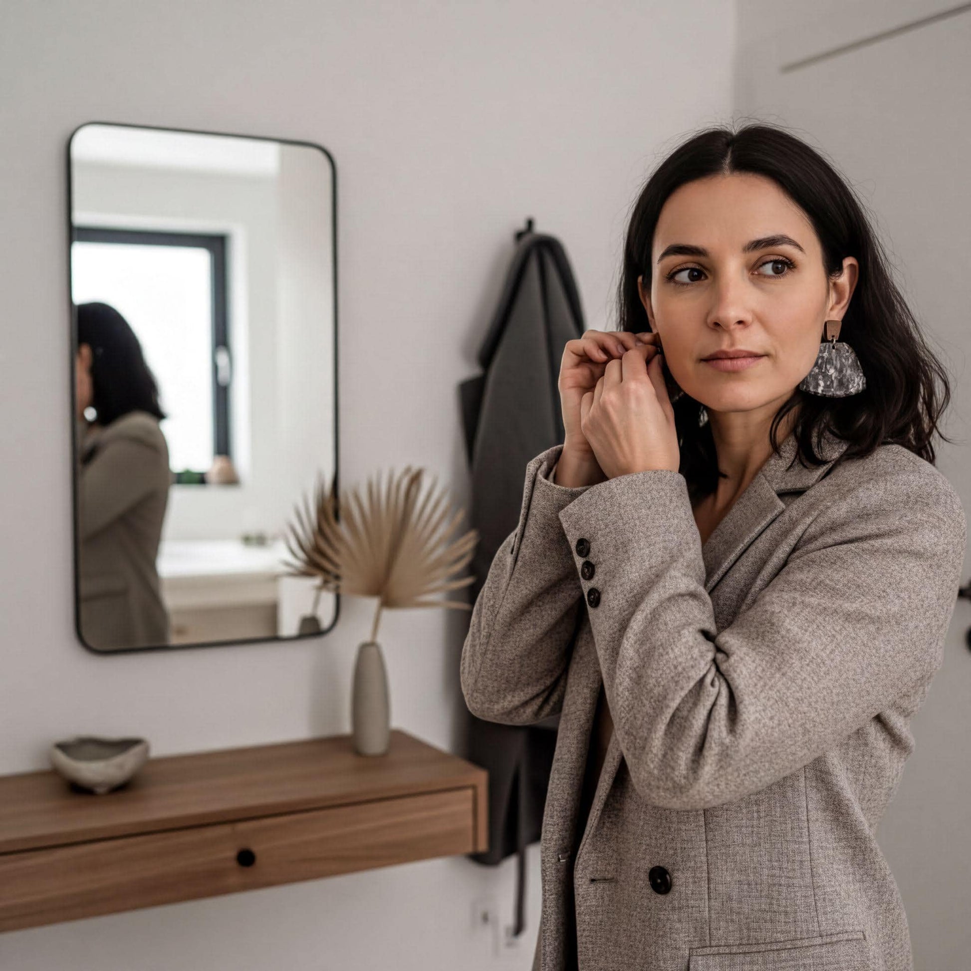 A woman in a gray blazer stands in a room, adjusting her large earrings. She is wearing a black jacket and has a serious expression on her face.