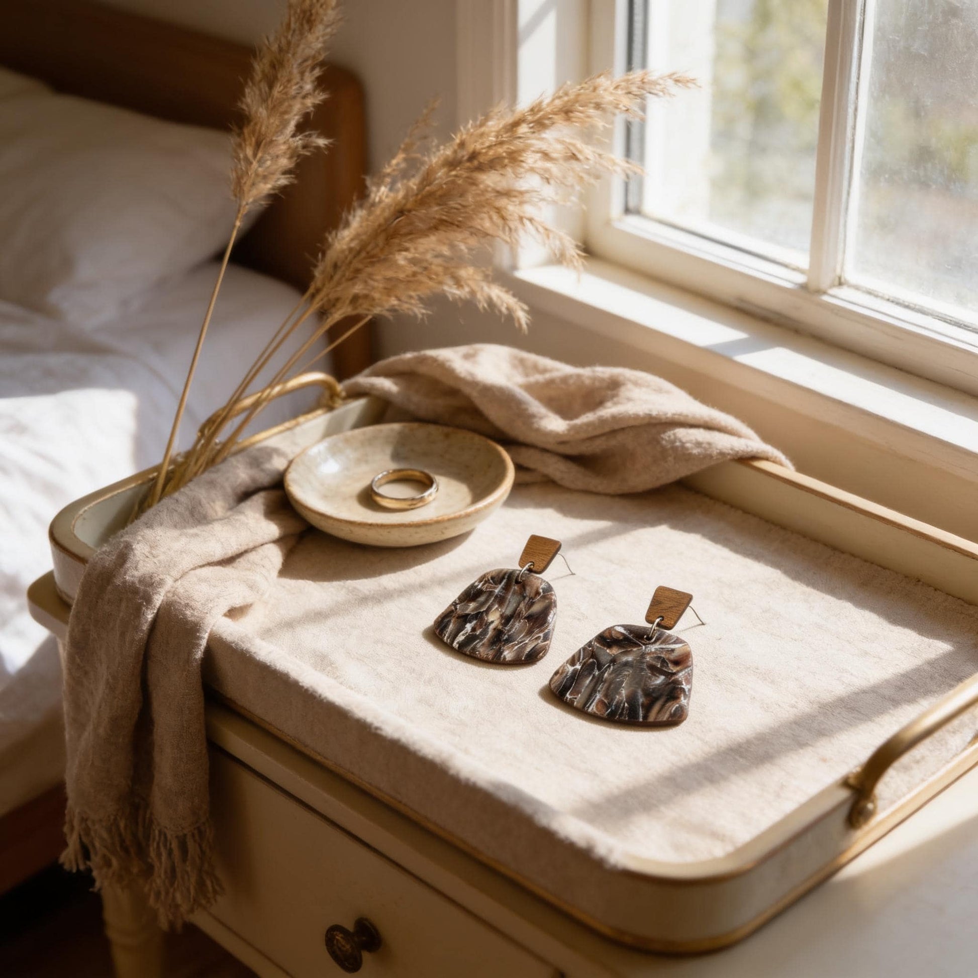 A tray with a pair of earrings, a ring, and a plant on a bedside table, with a window and a white blanket in the background.