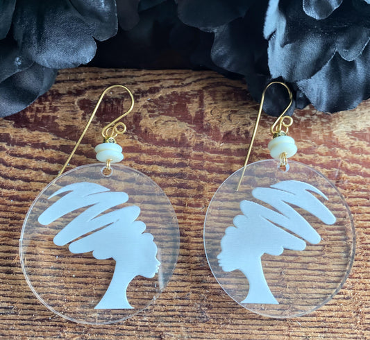 A pair of clear earrings with white silhouettes of a woman's head and hair designs on them, placed on a wooden surface.