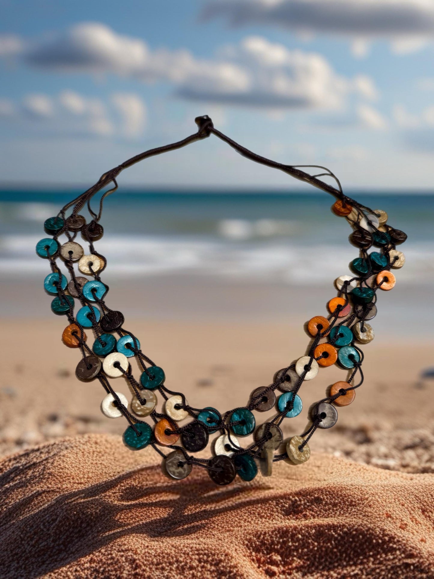 A colorful beaded necklace is displayed on a sandy beach, with the ocean and cloudy sky visible in the background.