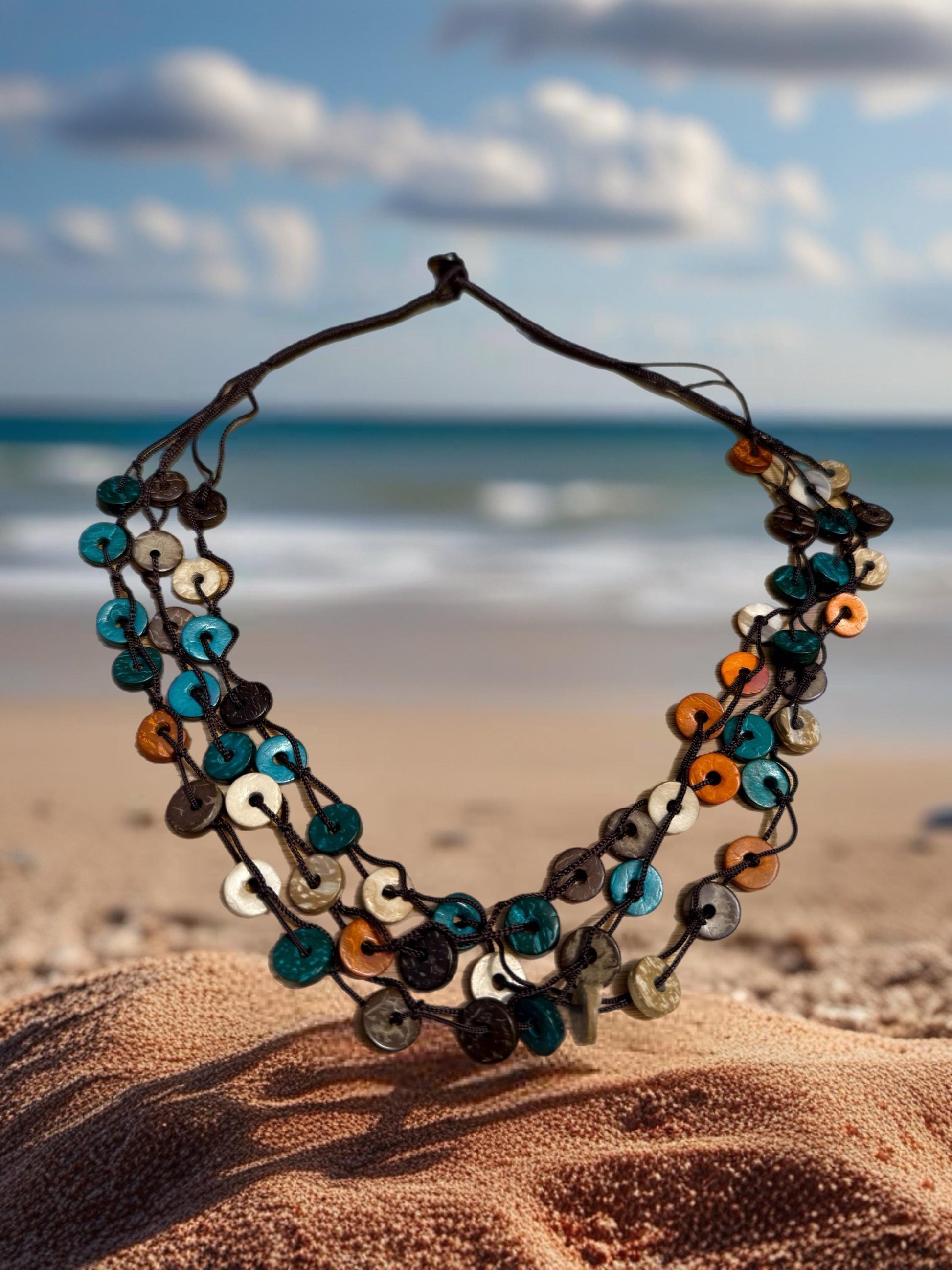 A colorful beaded necklace is displayed on a sandy beach, with the ocean and cloudy sky visible in the background.