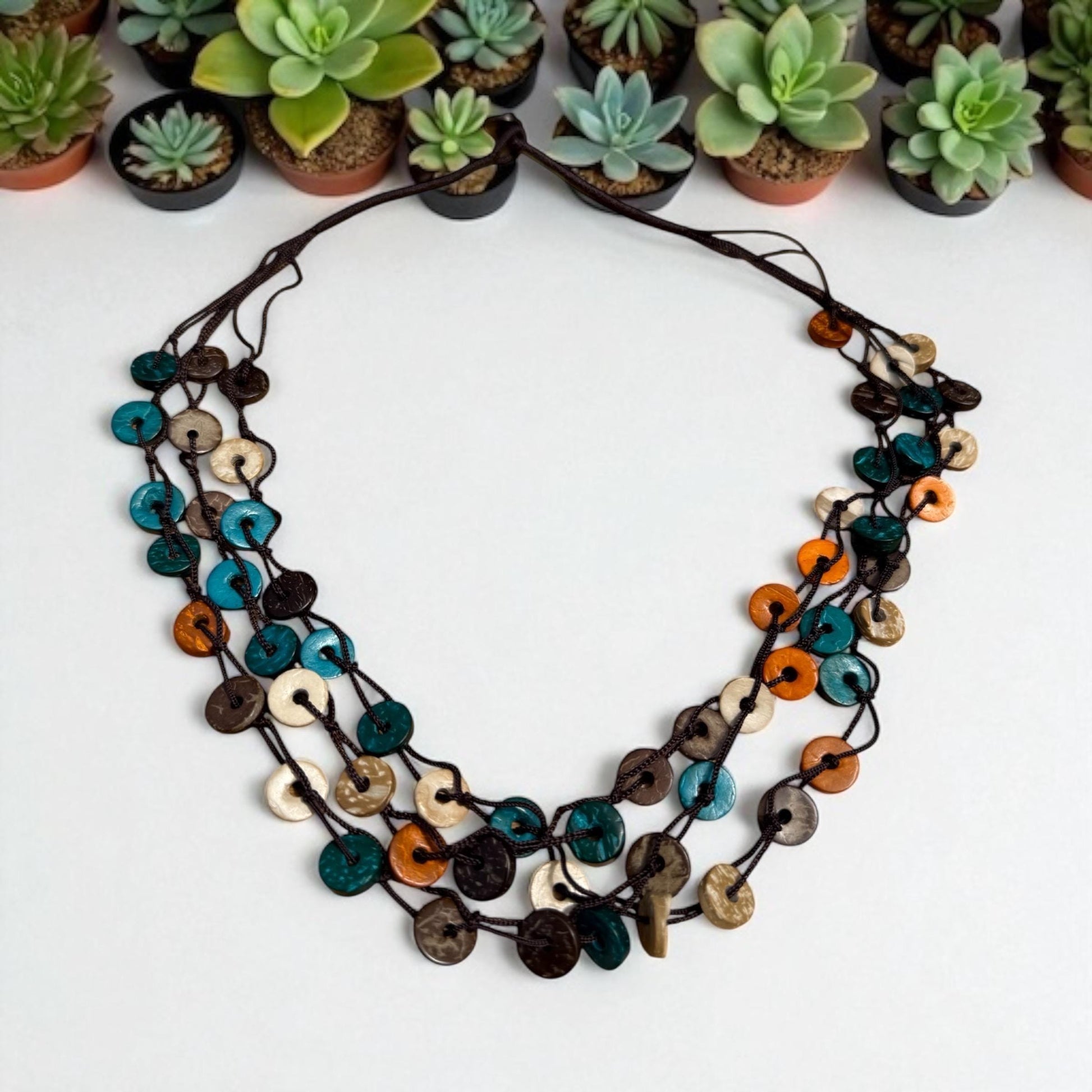 A colorful beaded necklace with various shades of blue, orange, and brown beads is displayed on a white surface, surrounded by potted succulents.