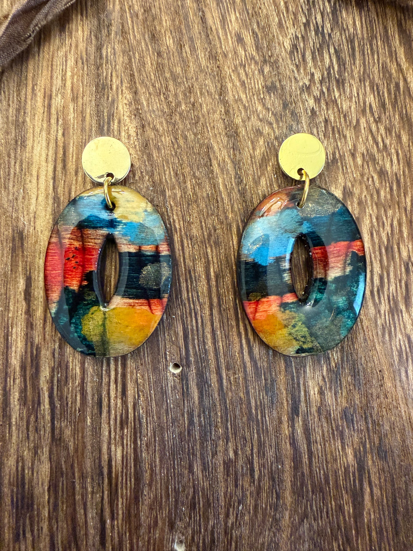A pair of colorful, oval-shaped earrings with gold-colored accents, displayed on a wooden surface.