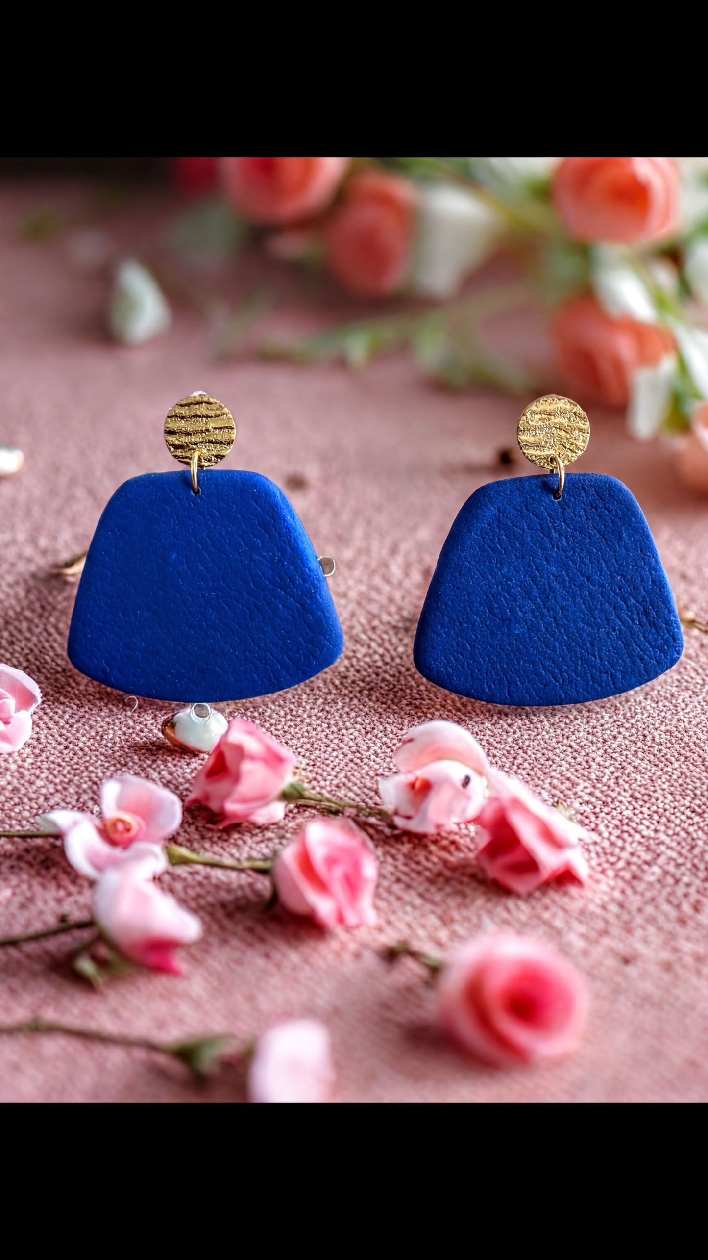 a pair of blue earrings with gold accents, placed on a pink surface with scattered pink flowers.