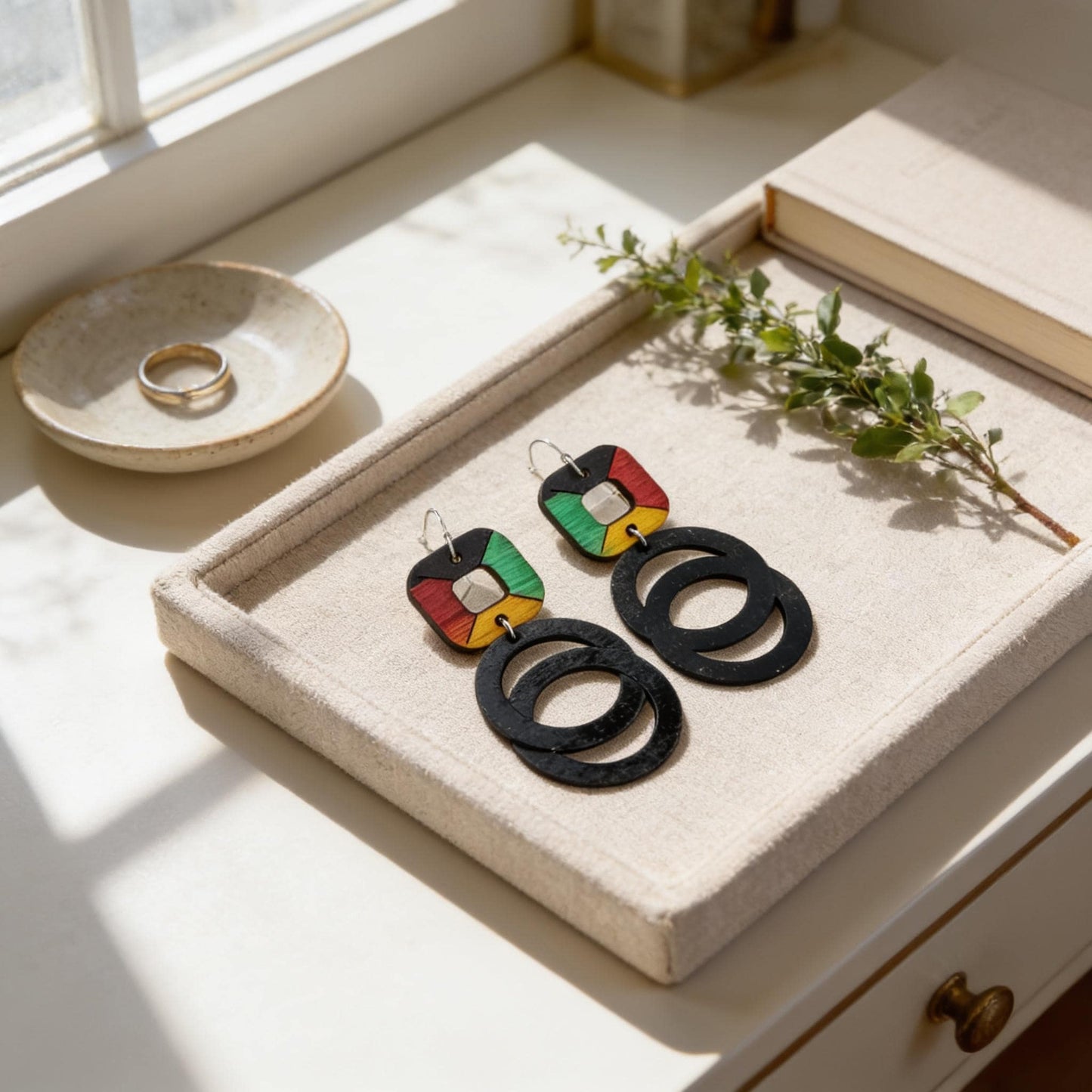 A pair of colorful earrings with black circular shapes rests on a beige tray, accompanied by a small white bowl and a plant branch.