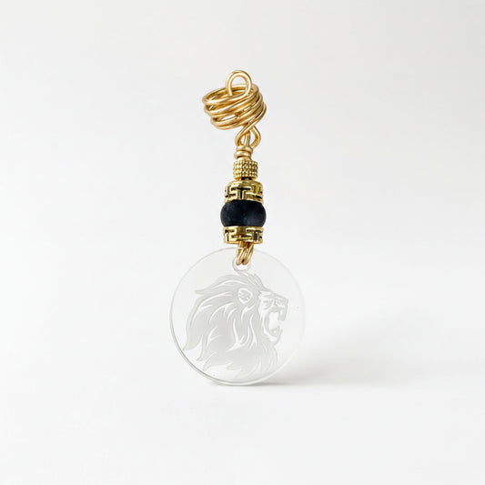 A clear pendant with a gold metal loop and a black bead, featuring a lion's head design.