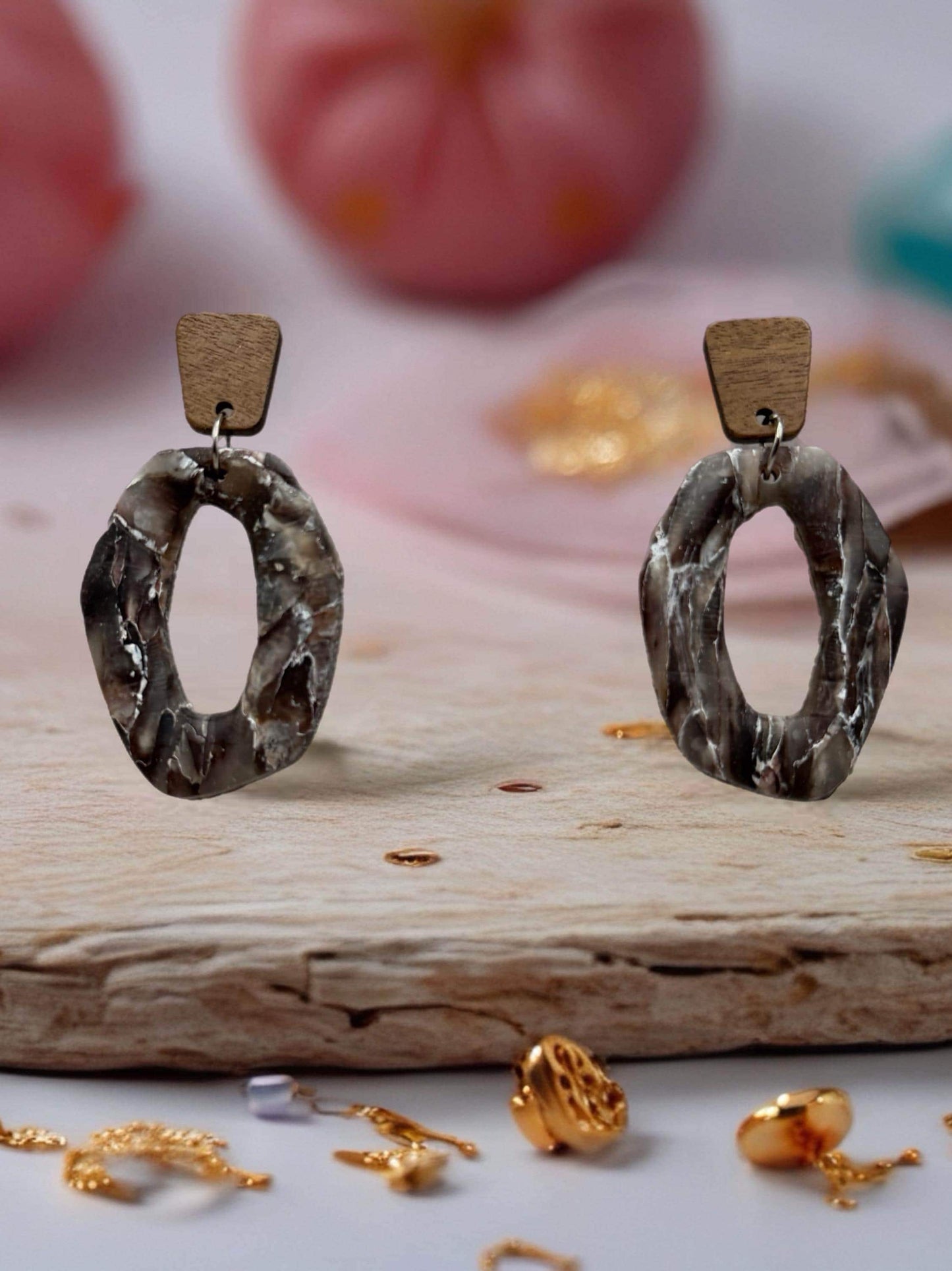a pair of earrings with a unique oval shape, placed on a wooden surface. The earrings are made of a material that resembles wood and have a metallic appearance. There are also some gold jewelry pieces scattered around the wooden surface, adding a touch of elegance to the scene.