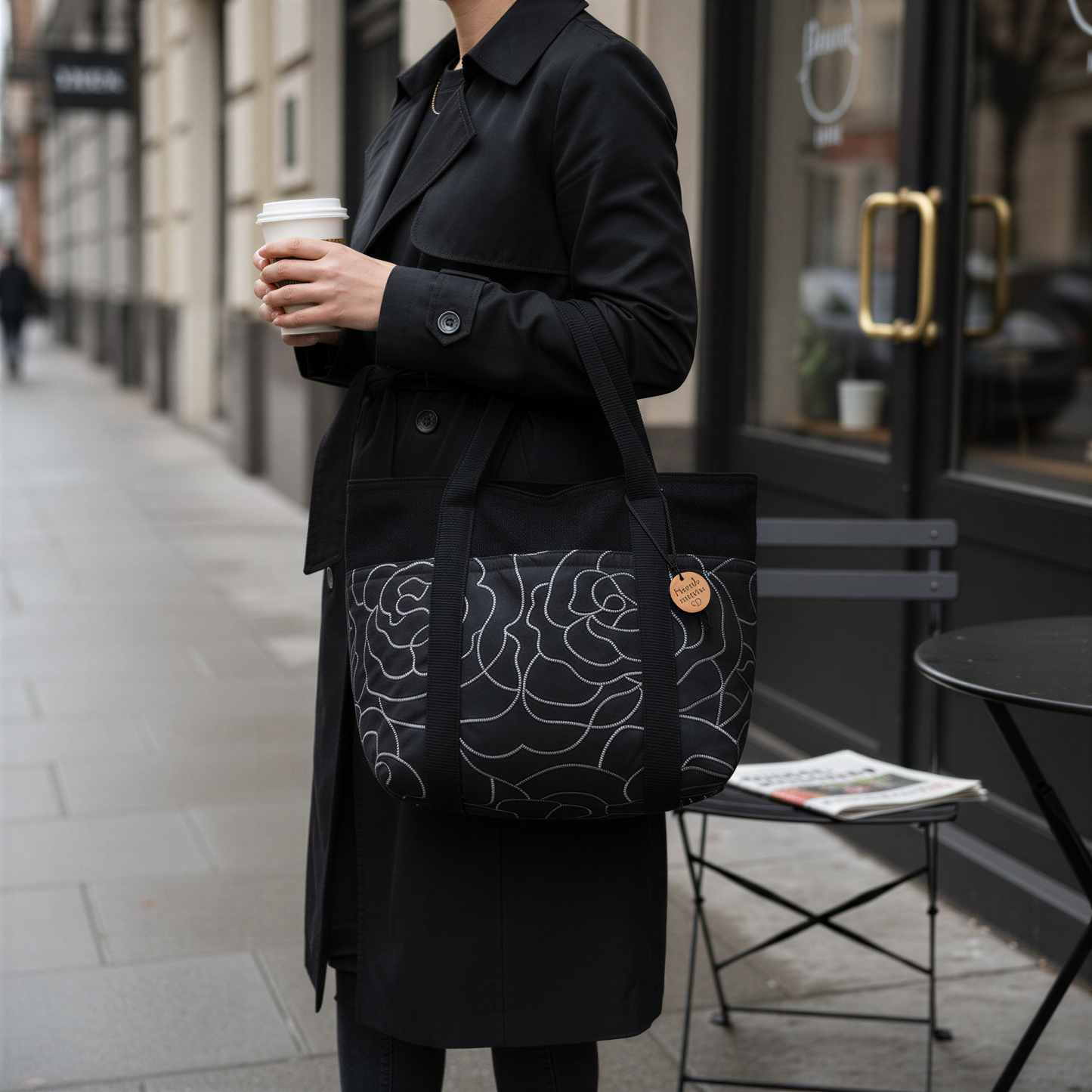 A person wearing a black coat and carrying a black and white floral tote bag is standing on a sidewalk, holding a coffee cup.