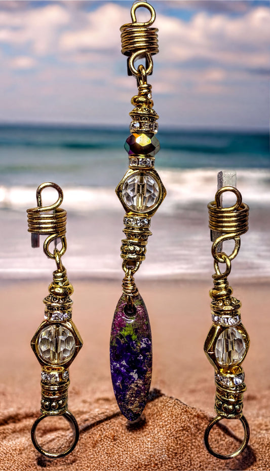 A set of three gold-colored earrings with various beads and charms, including a large purple and green stone pendant, displayed on a sandy beach with the ocean in the background.