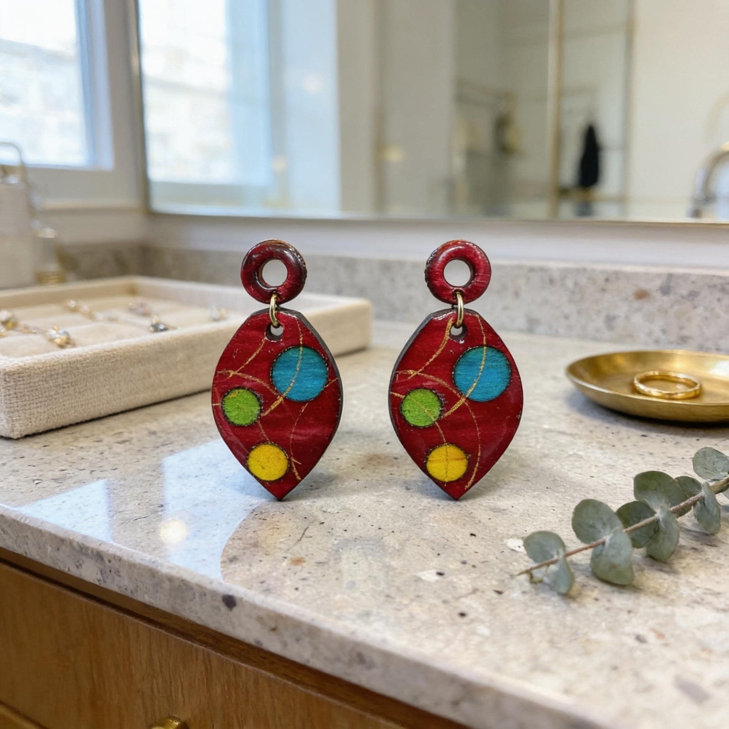 a pair of red earrings with colorful designs, placed on a countertop next to a small plant and a gold ring dish.